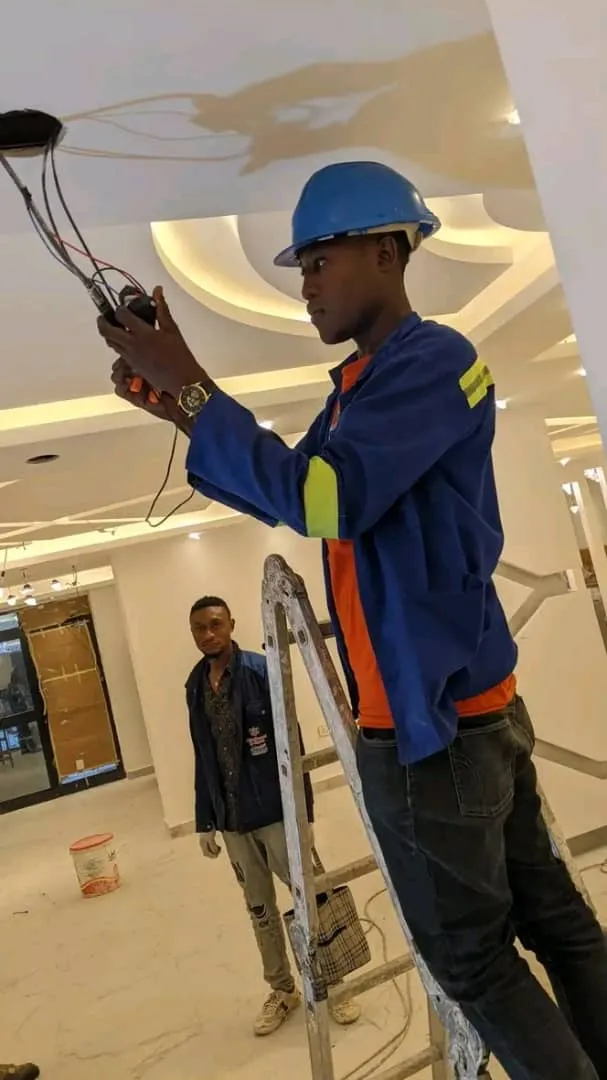 An electrician in a blue hard hat on a ladder wiring a ceiling light fitting, with a colleague looking on.